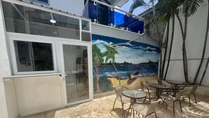 Un patio con un mural de una escena de playa, mobiliario exterior y un ventanal que da a un espacio interior.