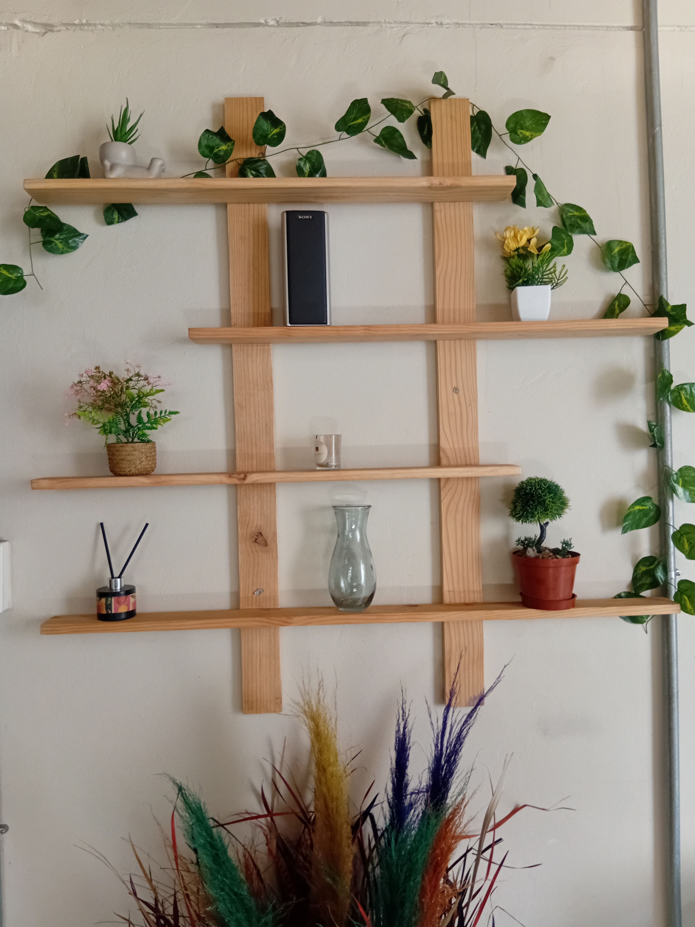 Una estantería de madera fijada a la pared, con varios objetos decorativos como plantas, un jarrón y un altavoz pequeño.