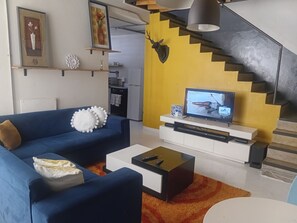 Un saló amb un sofà blau, una taula de centre, un televisor sobre un suport blanc i una escala amb una paret groga.