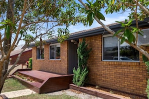 A brick house with a wooden deck, a small garden, and a tall evergreen tree. - Forster 8BR home, free parking, near shop & beach (Forster)