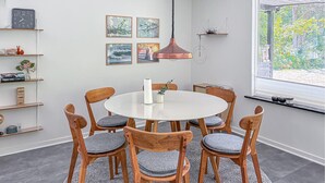 Ein Essbereich mit einem runden Tisch, sechs Holzstühlen mit grauen Kissen, einer hängenden Kupfer-Pendelleuchte und gerahmten Bildern an der Wand.