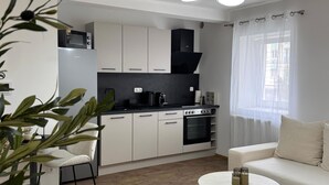 A modern kitchen with white cabinets, a black stove, and a microwave. There is a dining area with a white table and chairs, and a sofa with a white throw blanket.