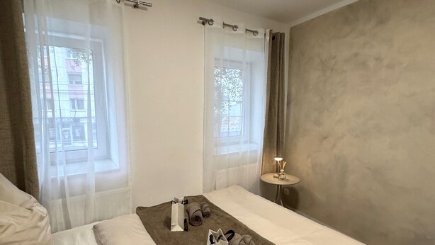A hotel room with two beds, a large window with sheer curtains, a bedside table with a lamp, and a brown throw blanket on one bed.