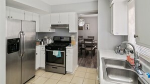 A modern kitchen with stainless steel appliances, a dining area with wooden chairs, and a large window with blinds. - Lovely 3BR Crystal Lake home Nearby park and Lake! (Crystal Lake)