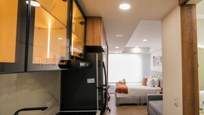A compact kitchen with a stove, refrigerator, and wooden cabinets. A bed and a sofa are visible in the background. - Tribeca Park by Jalo (Bogotá)