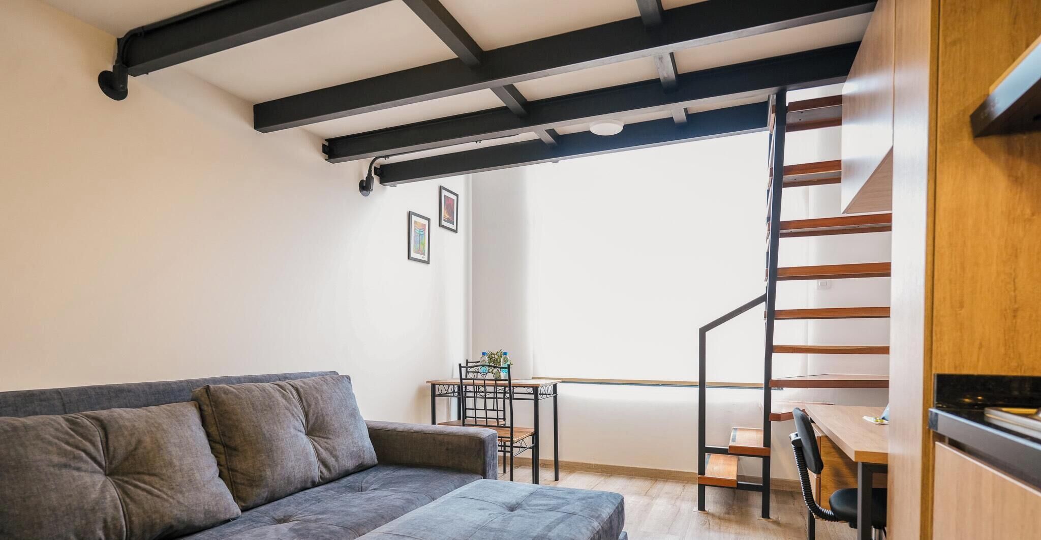 A modern living room with a grey sofa, a small table with chairs, a staircase, and a wooden cabinet.