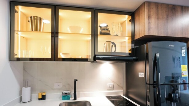 A modern kitchen with a black refrigerator, a built-in oven, and a microwave. There are cabinets with glass doors containing kitchenware. - Tribeca Park by Jalo (Bogotá)