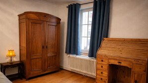 Quarto com armário de madeira, escrivaninha, radiador e janela com cortinas.