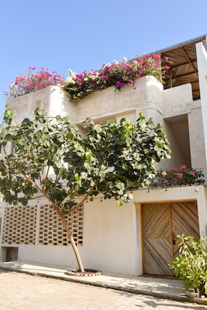 Ein Gebäude mit einem großen Baum davor, einem Balkon mit rosa Blüten und einer Holztür.