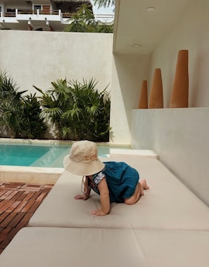Ein Kind krabbelt auf einem weißen Sofa im Freien in der Nähe eines Pools, im Hintergrund eine weiße Wand und Pflanzen.