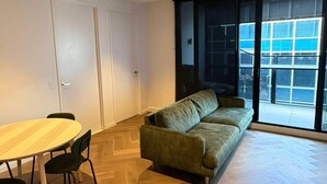 Un salón moderno con suelo de parqué de espiga, un sofá verde, una mesa de comedor con sillas y un ventanal con vistas a la ciudad.