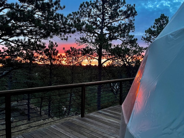 A wooden deck with a railing, overlooking a forested area with a sunset in the background.