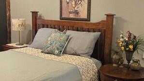 A bedroom with a wooden bed, a nightstand with a lamp, a framed floral painting on the wall, and a small table with decorative items.