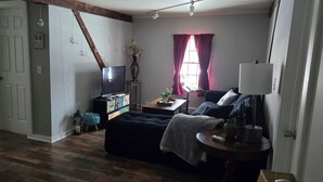 A living room with a sofa, a television, a small table with a lamp, and a window with curtains.