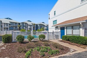 A multi-story residential building with a white exterior, a blue door, and a fenced courtyard with small trees and shrubs. - Costa Vista Townhouse #13 (Miramar Beach)