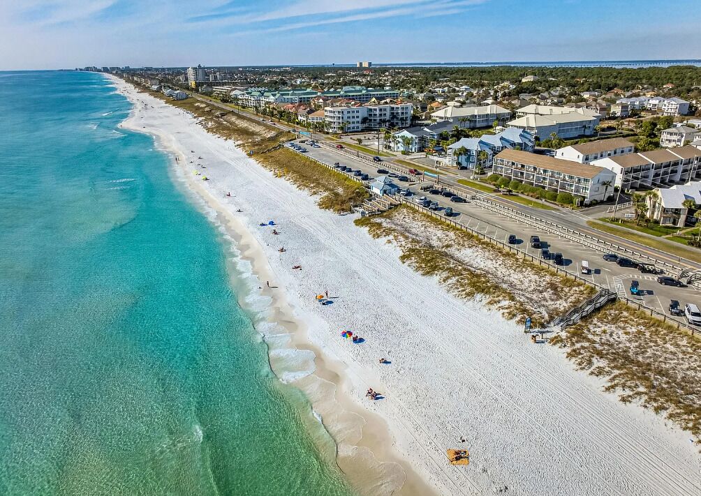 An aerial view of a beachside area with a long stretch of white sand, clear turquoise water, and a row of buildings along the shore.