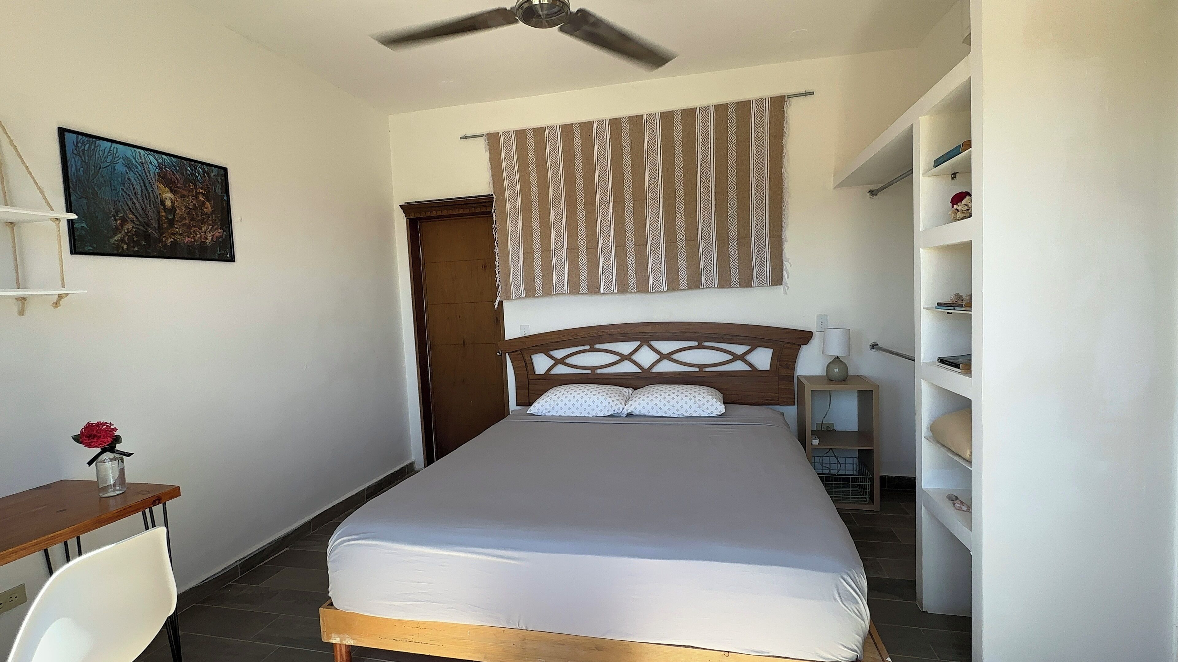 Une chambre à coucher comprenant un lit, un bureau, une chaise, un ventilateur de plafond, une étagère avec des objets décoratifs et un rideau à motifs.