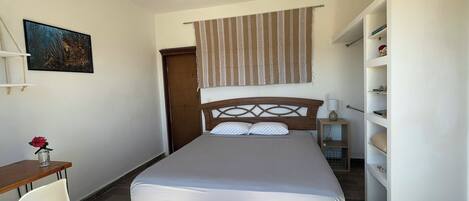 Une chambre à coucher comprenant un lit, un bureau, une chaise, un ventilateur de plafond, une étagère avec des objets décoratifs et un rideau à motifs.
