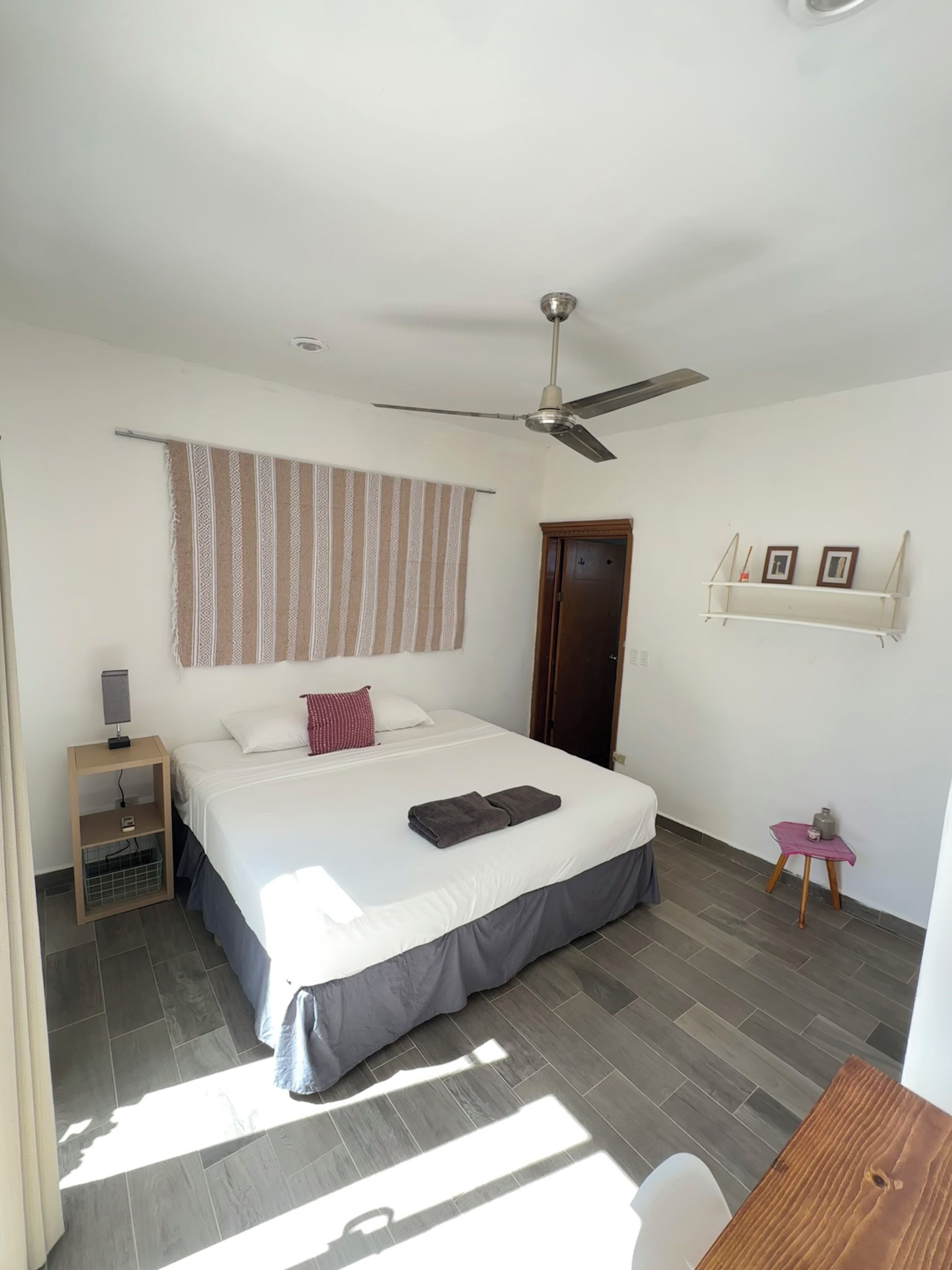 Une chambre à coucher avec un lit, un ventilateur de plafond, une table de chevet, une petite table et une porte.
