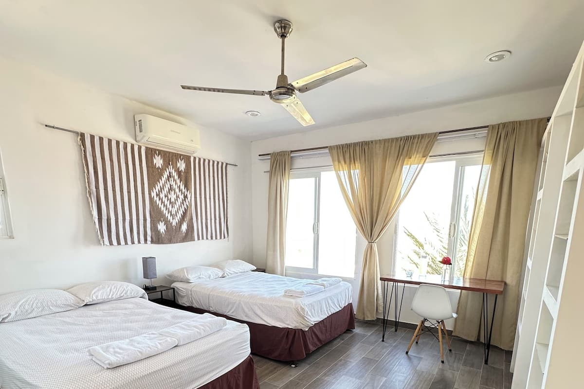 Een hotelkamer met twee bedden, een plafondventilator, een airconditioning, een bureau met stoel en een groot raam met gordijnen.