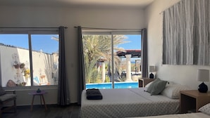 A hotel room with a large sliding glass door leading to a pool area, a bed with a patterned bedspread, a bedside table with a lamp, and a chair with a small table. - The Colony La Ventana (La Ventana)