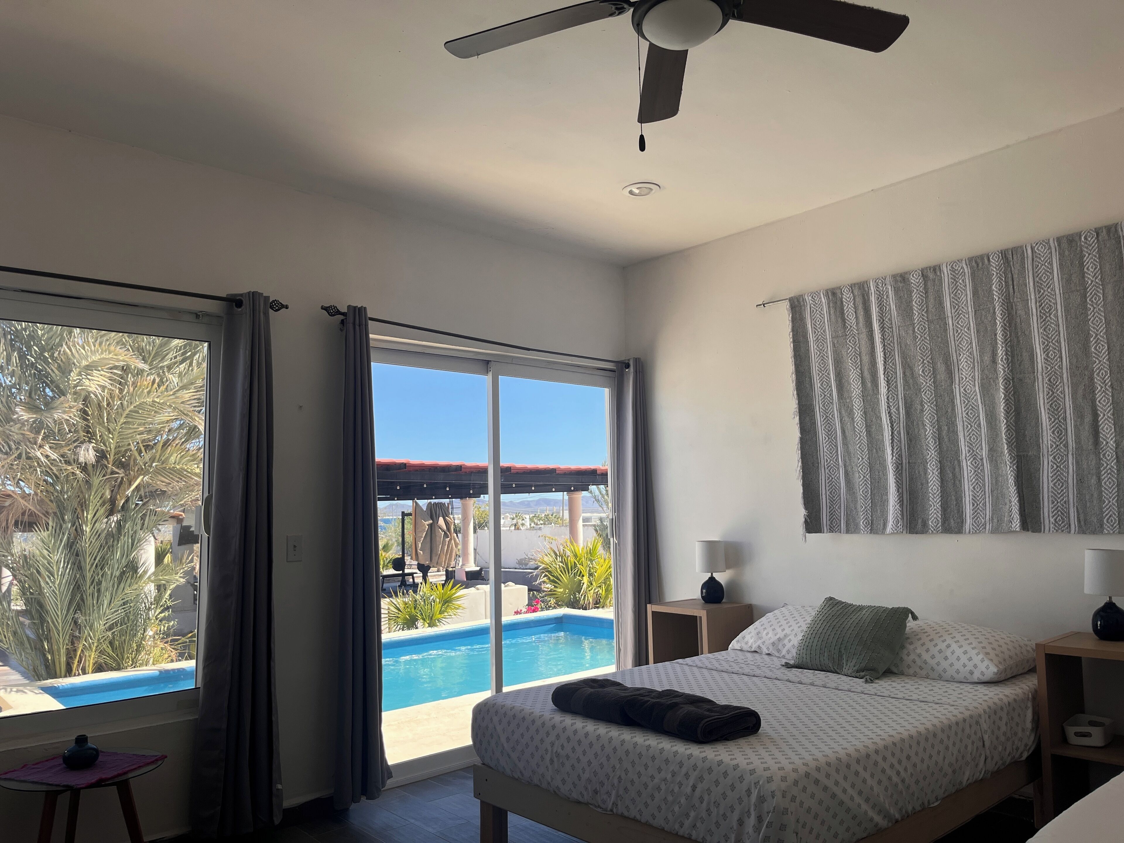Ein Schlafzimmer mit Bett, Nachttisch, Lampe, Deckenventilator und Blick auf einen Swimmingpool und Palmen durch eine Glastür.