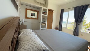 A bedroom with a bed, a wooden headboard, a bedside table with a lamp, a wardrobe with shelves, and a window with curtains. - The Colony La Ventana (La Ventana)