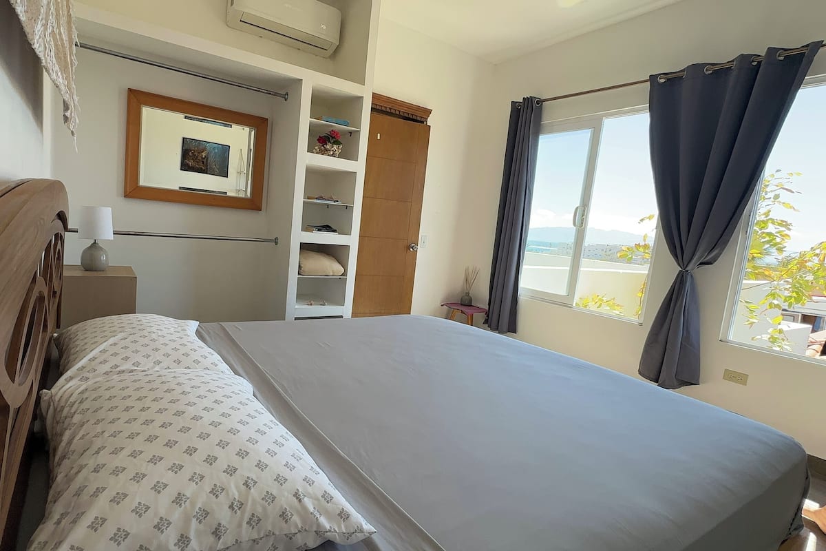 Een slaapkamer met een bed, een houten hoofdbord, een nachtkastje met een lamp, een inloopkast met planken en een raam met gordijnen.