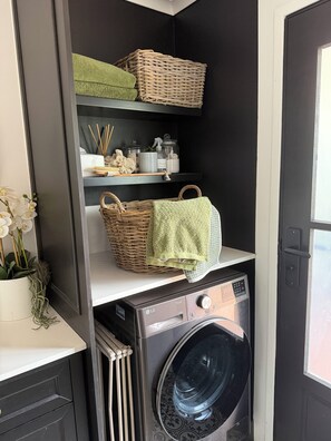 A laundry area with a washing machine, wicker baskets, and shelves with storage items. - Adorable one-bedroom studio with a cozy fire pit (Glenbrook)