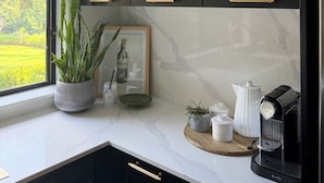 A kitchen with dark cabinets, a marble countertop, a coffee machine, and a window with a view of greenery.