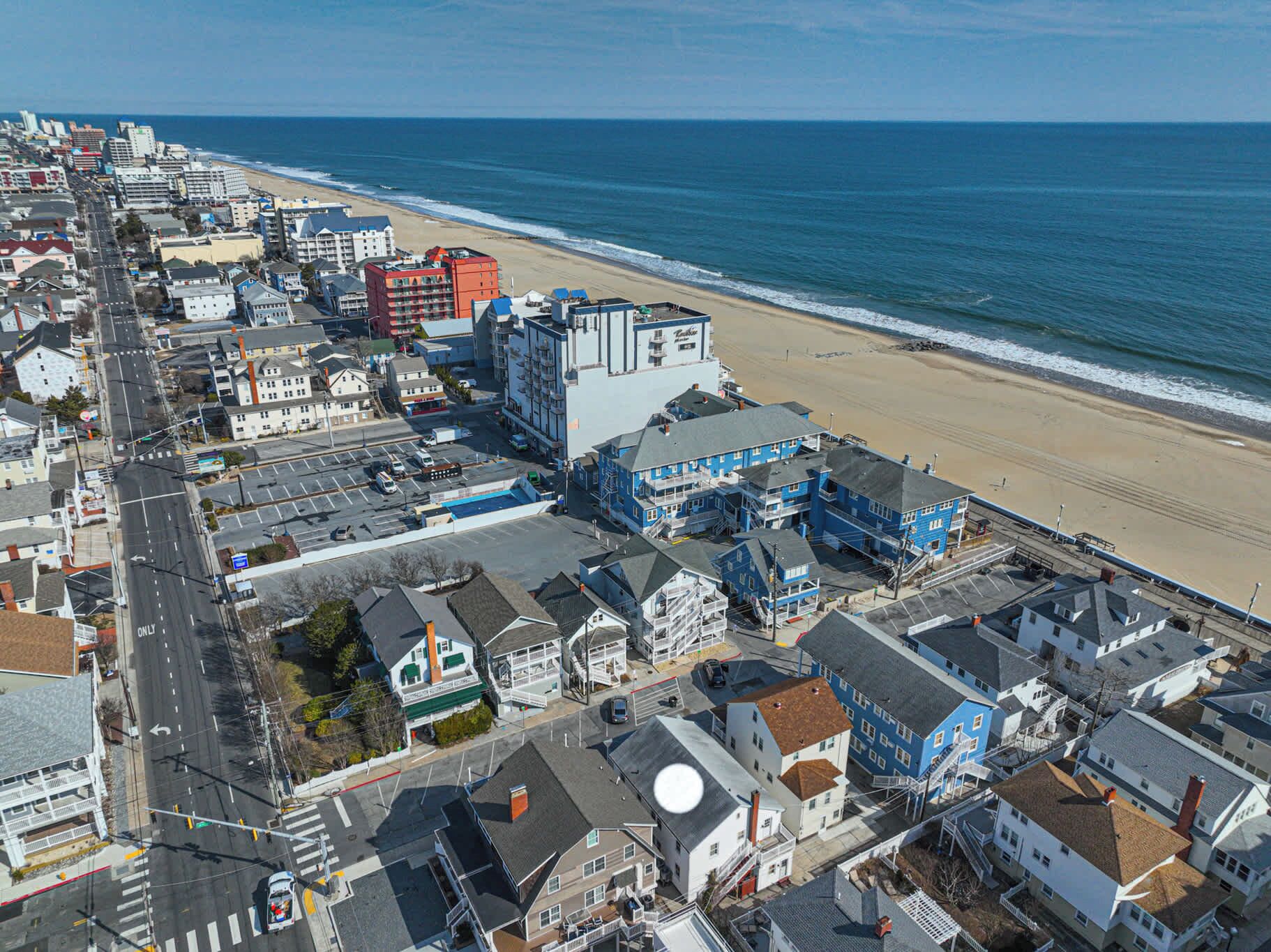 An aerial view of a coastal town with a beach, buildings, and a prominent hotel with multiple floors and balconies.