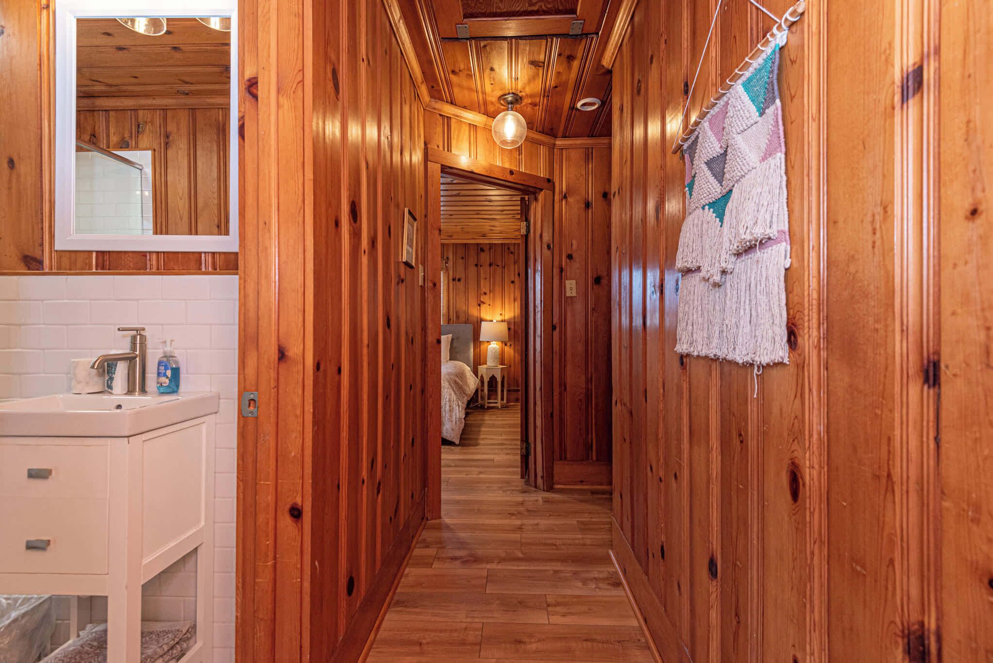 A wooden hallway with a sink and mirror on the left, a bed with a lamp in the distance, and a hanging macrame piece.
