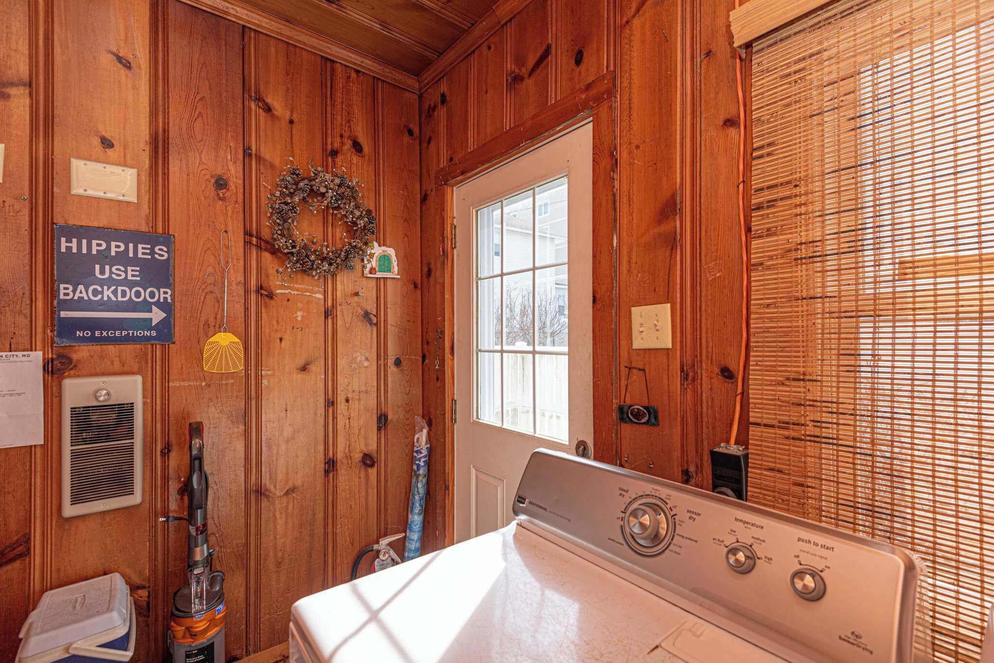 A laundry room with a washer and dryer, a sign indicating 'HIPPIES USE BACKDOOR NO EXCEPTIONS', and a wreath on the door.