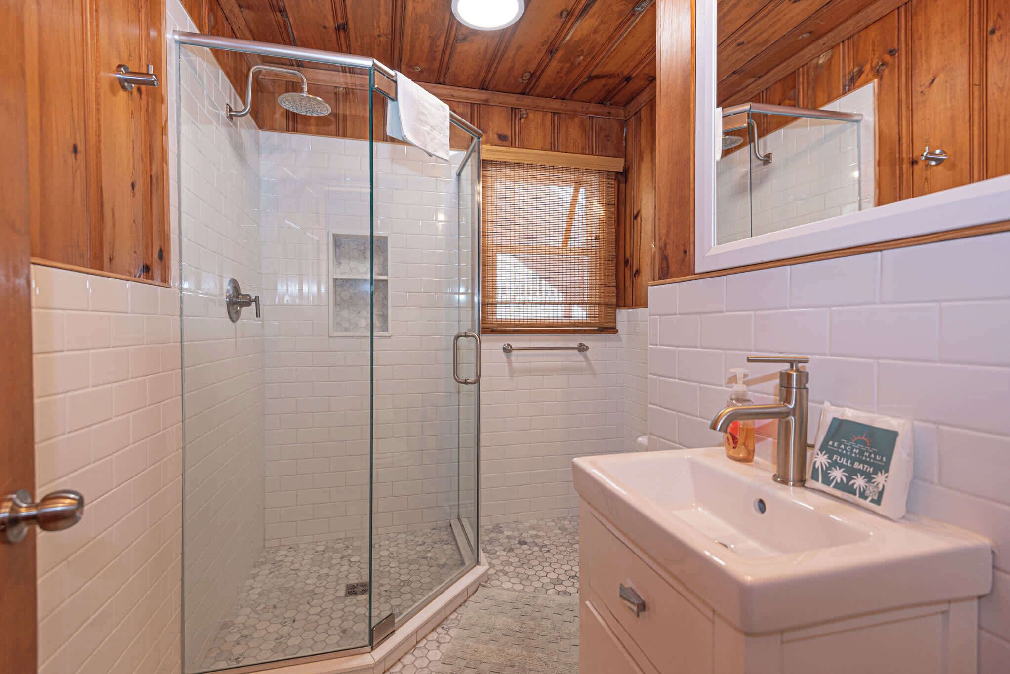 Un baño con ducha envuelta en vidrio, paredes de azulejos blancos, armarios de madera y un lavabo con dispensador de jabón.