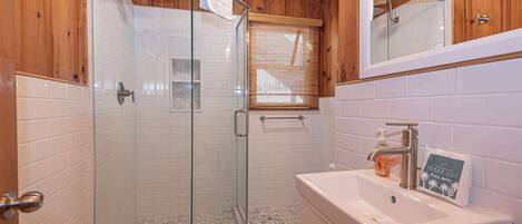 Un baño con ducha envuelta en vidrio, paredes de azulejos blancos, armarios de madera y un lavabo con dispensador de jabón.