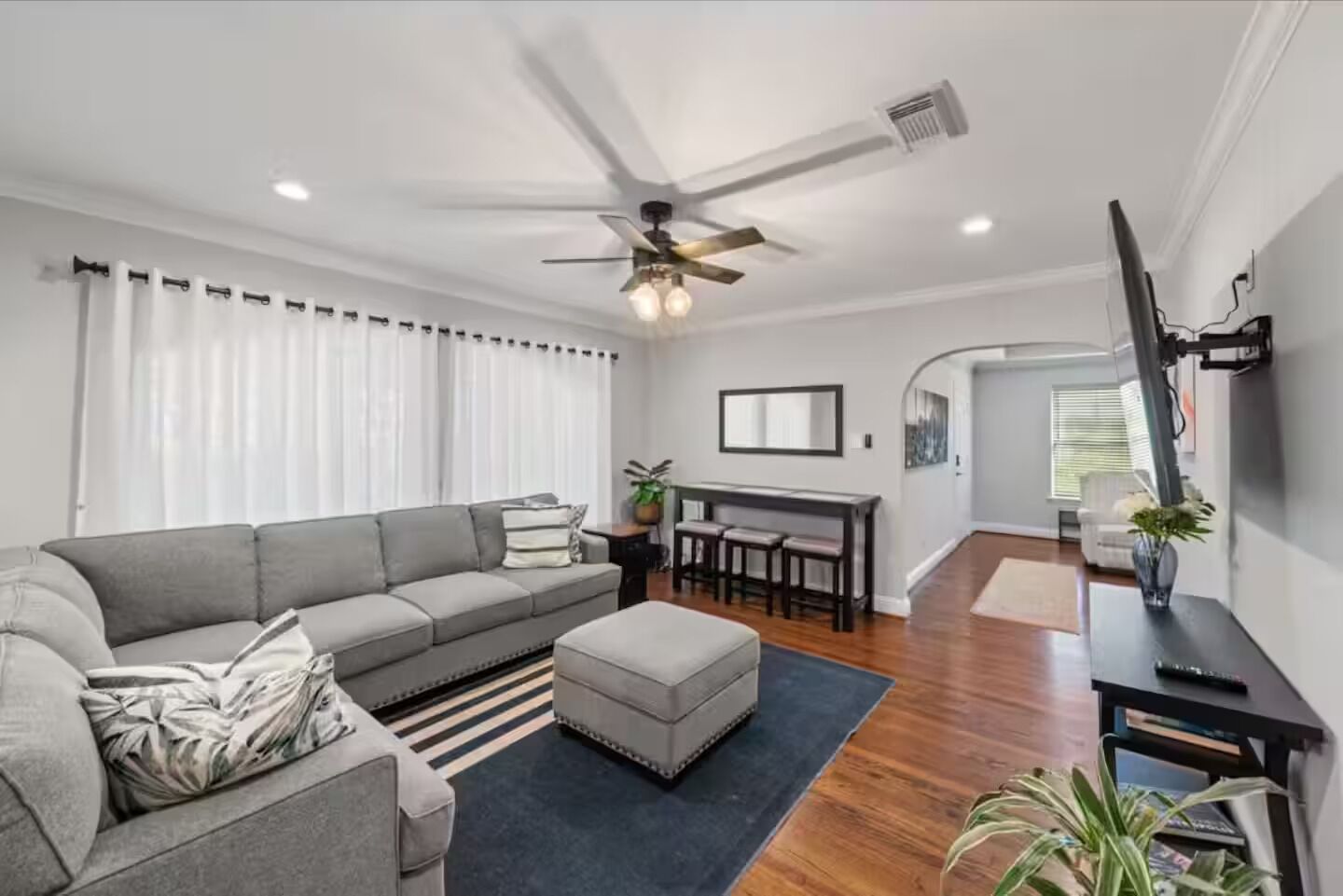 Ein modernes Wohnzimmer mit grauem Sofa, Couchtisch und Deckenventilator. Ein Essbereich mit Tisch und Stühlen, sowie ein Flur, der in einen anderen Raum führt.