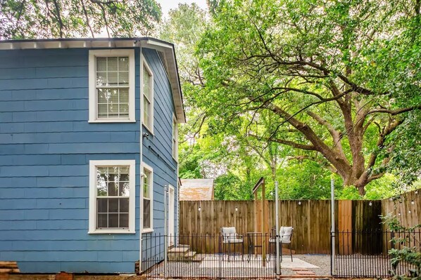 Ein blaues Haus mit einem umzäunten Garten, einem großen Baum und Sitzgelegenheiten im Freien.