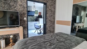 A bedroom with a large bed, a TV on a wooden stand, and a desk with a chair. There is a door leading to a kitchenette and a bathroom. - Nice little apartment (Pornic)
