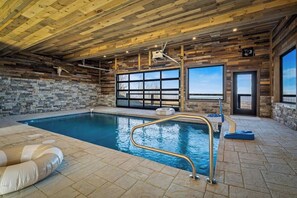 An indoor swimming pool with a wooden ceiling and stone walls, a clear view outside, and a poolside area with a lounge chair and inflatable ring. - 5-Bedroom Cabin with Indoor Pool, Pickleball Court & Hot Tub Near Dollywood (Sevierville)