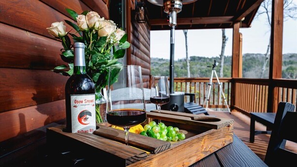 Une table en bois, une bouteille de vin, des verres et un bouquet de roses sont disposés sur une terrasse offrant une vue sur l’extérieur.