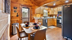L’intérieur d’un chalet en bois comprend une salle à manger avec une table dressée pour quatre personnes, une cuisine avec des meubles en bois, un réfrigérateur et une poubelle.