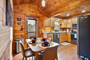A wooden cabin interior with a dining table set for four, a kitchen with wooden cabinets, a refrigerator, and a trash can. - Twig Hollow-Hot tub, fire pit, dog friendly, view (Mineral Bluff)