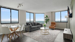 Ein geräumiges Wohnzimmer mit grauem Sofa, weißen Stühlen, einem runden Couchtisch und einem Esstisch mit Stühlen. Große Fenster bieten Blick auf die Stadt.