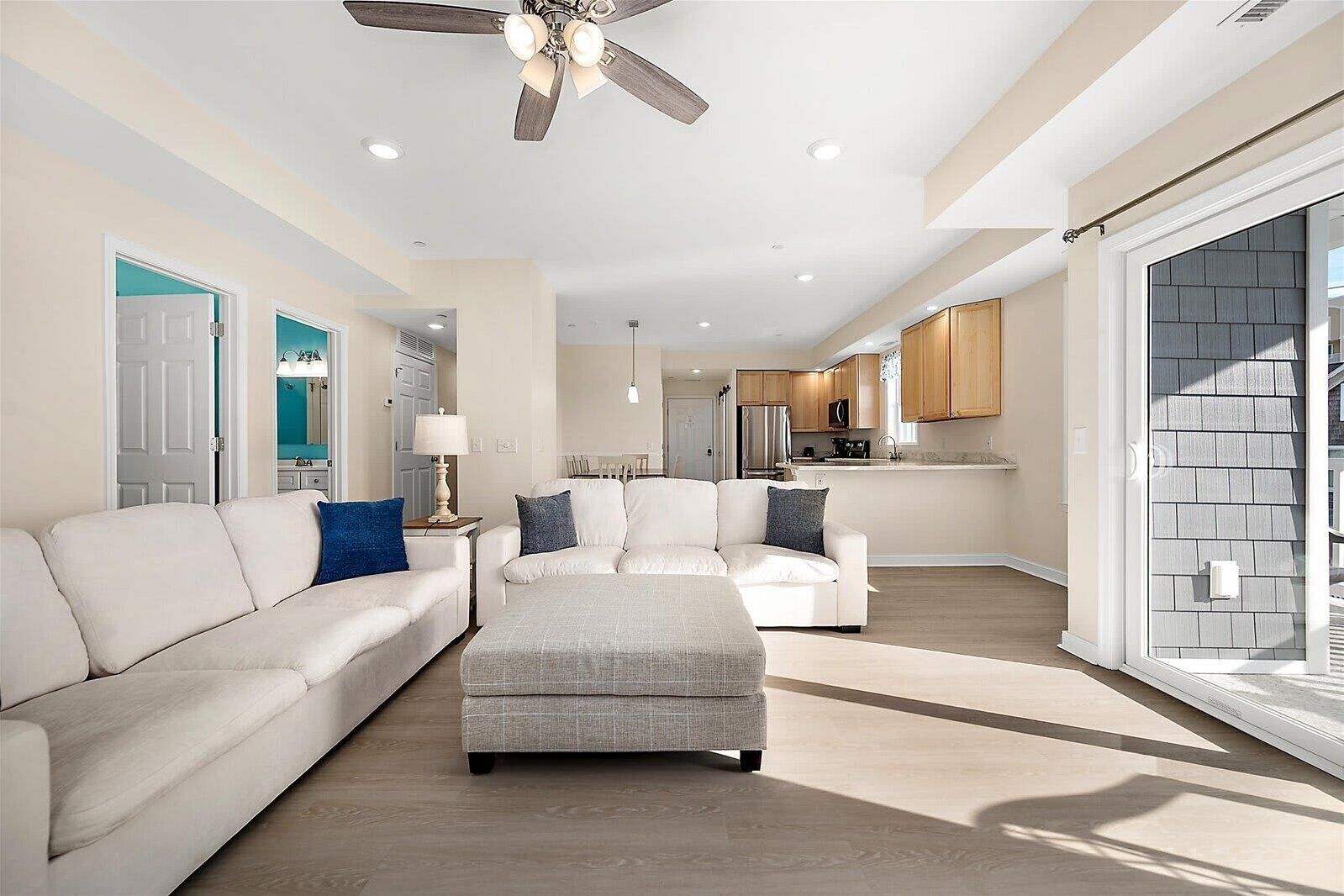 A spacious living room with a large white sofa, a grey ottoman, and a ceiling fan.