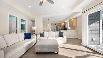 A spacious living room with a large white sofa, a grey ottoman, and a ceiling fan.
