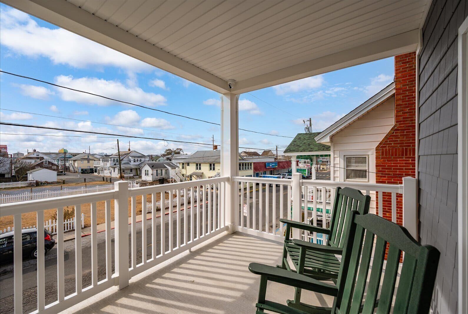 A covered porch with a white railing, green chairs, and a view of a street with houses and a gas station.