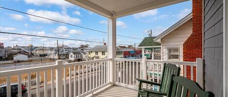 A covered porch with a white railing, green chairs, and a view of a street with houses and a gas station.