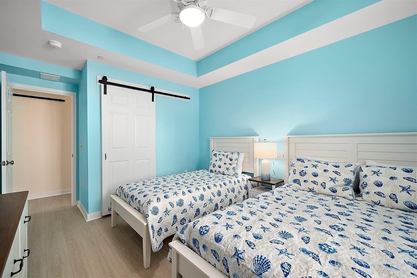 A bedroom with two beds, a ceiling fan, a nightstand with a lamp, and a door.