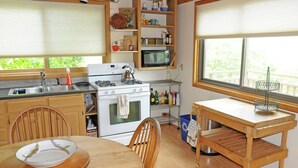 A compact kitchen with a stove, microwave, and sink. There is a dining table with chairs, and a window with blinds.