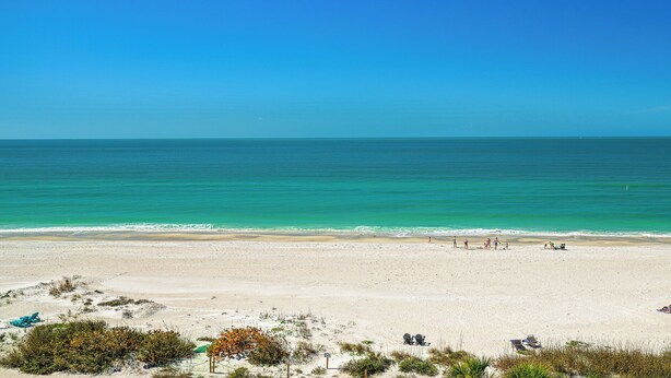 Uma extensa praia de areia dourada com águas turquesa cristalinas, algumas pessoas e espreguiçadeiras.